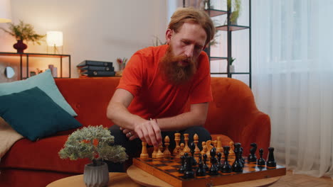 Focused-bearded-redhead-young-man-playing-chess-leisure-board-game-alone,-domestic-activity-at-home