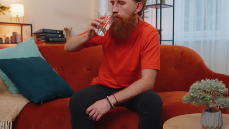 Thirsty-young-adult-man-sitting-at-home-holding-glass-of-natural-aqua-make-sips-drinking-still-water