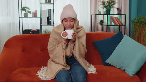 Sick-young-woman-wear-hat-wrapped-in-plaid-sit-alone-shivering-from-cold-on-sofa-drinking-hot-tea