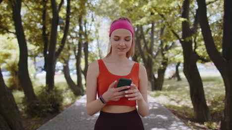 Athletic-fitness-sport-runner-girl-walking,-using-smartphone-listening-music-with-earphones-in-park