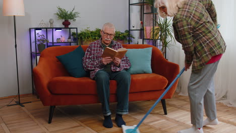 Senior-grandmother-cleans-wipes-dust-on-floor-with-mop-under-grandfather-legs-while-he-reading-book