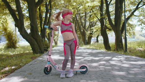 Athletic-fit-young-girl-rides-on-electric-scooter,-standing-near-bike-looking-at-camera-in-park