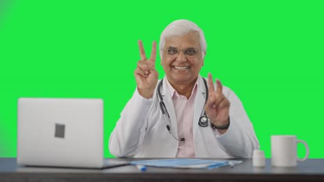 Happy-Indian-senior-doctor-showing-victory-sign-Green-screen