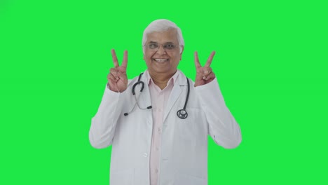 Happy-Indian-senior-doctor-showing-victory-sign-Green-screen