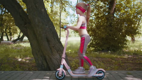 Young-athletic-fit-blonde-girl-rides-traveling-on-electric-scooter-on-road-in-park-on-sunny-day