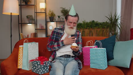 Happy-senior-old-man-celebrating-birthday-party,-makes-wish-blowing-burning-candle-on-small-cupcake