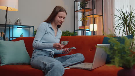 Smiling-happy-girl-counting-money-cash-and-use-laptop-pc-calculate-domestic-income-earnings-at-home