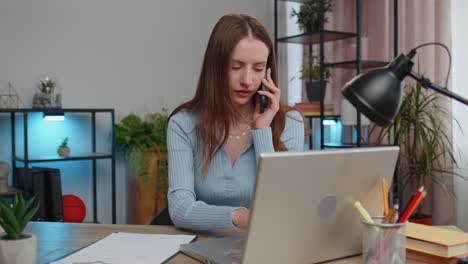 Young-woman-freelancer-answering-to-client-on-mobile-phone-call,-using-laptop-computer,-home-office