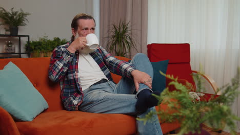 Attractive-smiling-senior-old-grandfather-man-drinking-a-cup-of-coffee-or-herbal-tea-at-home-couch