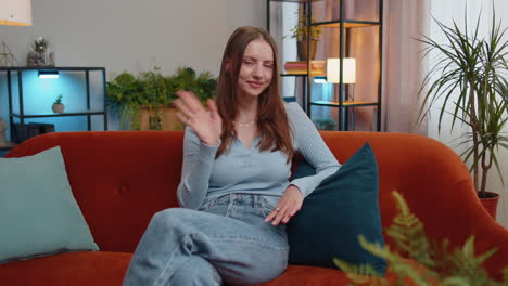 Young-woman-smiling-friendly-at-camera,-waving-hands-gesturing-hello,-hi,-greeting-at-home-on-couch