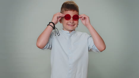 Seductive-cheerful-toddler-stylish-boy-child-in-shirt-wearing-sunglasses-charming-smile-on-blue-wall