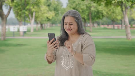 Cheerful-Indian-old-woman-scrolling-through-phone-in-park