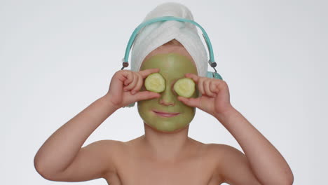 Smiling-child-girl-moisturizing-green-cucumber-mask-on-face-listening-to-music-on-headphones-dancing