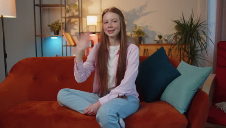 Child-girl-smiling-friendly-at-camera,-waving-hands-gesturing-hello,-hi,-greeting-at-home-on-couch