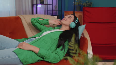 Happy-young-woman-in-wireless-headphones-relaxing-lying-on-sofa-at-home-listening-favorite-music