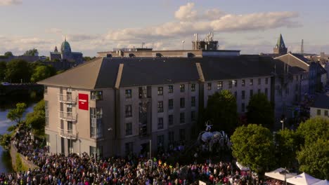 Busy-Galway-city-center-during-the-Pegasus-Parade,-with-crowds-following-the-procession-to-Spanish-Arch,-Ascending-aerial-shot