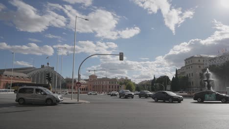 Vista-Del-Tráfico-Con-La-Fuente-De-La-Alcachofa-Y-La-Estación-De-Atocha-De-Fondo,-Madrid,-España