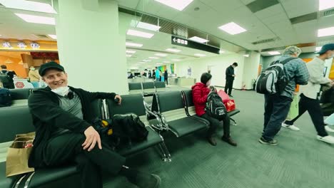 White-caucasian-man-with-a-cap-and-face-mask-on-chin-sits-and-waits-to-board-at-an-Asian-airport-gate,-surrounded-by-other-travelers