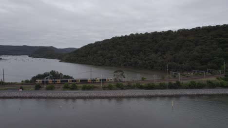 Train-in-countryside-shot-on-drone-with-river-and-trees-surrounding-on-a-gloomy-day