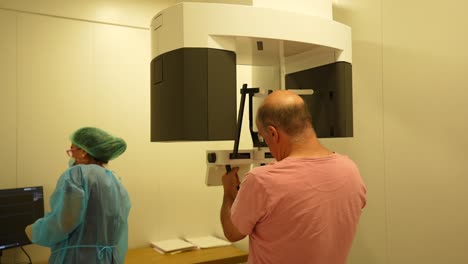 Rear-shot-of-female-dentist-with-male-patient-trying-to-place-his-head-in-dental-x-ray-machine