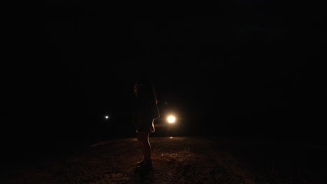 Woman-in-a-dress-is-dancing-alone-in-the-dark-with-only-car-lights-in-the-background-at-night,-copy-space