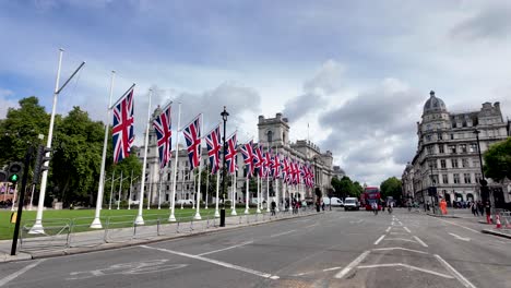 Fila-De-Banderas-De-La-Union-Jack-Que-Adornan-Los-Jardines-De-Parliament-Square-En-Westminster,-Londres,-Con-Edificios-Históricos-Al-Fondo