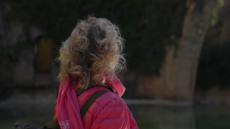 A-person-with-curly-blond-hair-is-seen-from-behind-wearing-a-pink-jacket,-standing-in-an-outdoor-setting-with-greenery-and-a-large-stone-arch-in-the-background,-capturing-a-serene-moment-in-nature