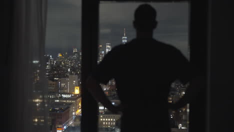 Man-stands-next-to-apartment-or-office-or-hotel-window-and-opens-curtain-to-amazing-cityscape-view-with-skyscrapers-at-night