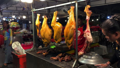 Cerdo-Asado-Glaseado-Cuelga-De-Ganchos-En-Un-Marco-De-Metal-En-Un-Mercado-Nocturno-Asiático-Mientras-El-Vendedor-Revuelve-La-Sopa-Y-Los-Niños-Pasan-Caminando