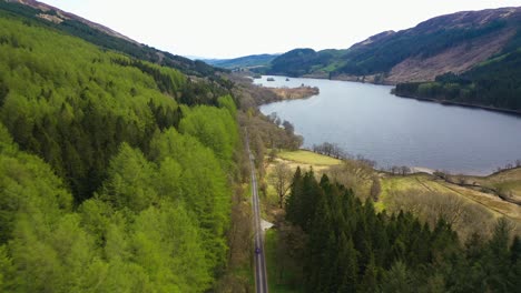 Aerial-view-of-car-driving-along-scenic-road-through-green-pine-tree-forest-on-Loch-Chon-in-the-Scottish-highlands,-Scotland