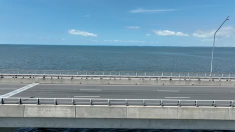 Vista-Lateral-Del-Dron-En-Una-Toma-De-Seguimiento-Del-Puente-Conmemorativo-Ted-Smout,-Cámara-Sobre-El-Agua-En-La-Bahía-De-Moreton-Con-El-Océano-Abierto-De-Fondo,-Tomada-Al-Mediodía-En-Un-Día-Claro-Y-Soleado