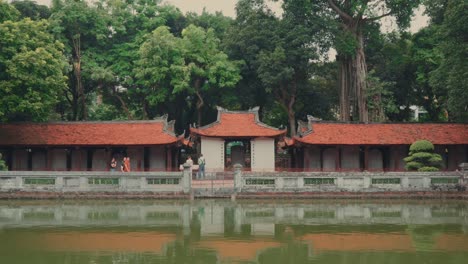 People-walk-in-front-of-green-lake-at-the-temple-of-literature-pagoda-background,-golden-hour