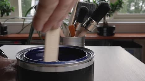 Men's-hands-mixed-blue-paint-with-a-wooden-stick-on-a-wooden-table-with-painting-brushes-on-a-background