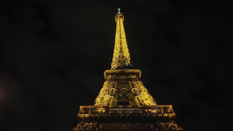 La-Parte-Superior-De-La-Torre-Eiffel-Iluminada-Con-El-Símbolo-De-Los-Anillos-Olímpicos-Antes-De-Los-Juegos-De-2024-Por-La-Noche-En-El-Centro-De-París,-Francia.