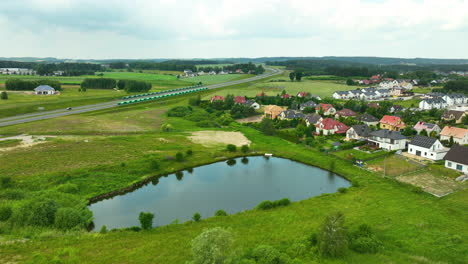 Vista-Aérea-De-Un-Barrio-Suburbano-Con-Un-Pequeño-Estanque-Y-Campos-Verdes-Que-Lo-Rodean.