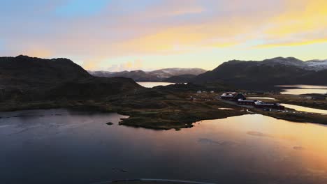 Toma-Aérea-De-La-Naturaleza-Noruega-Con-Un-Lago,-Montañas-Y-Una-Barraca-De-Madera-Al-Fondo-Al-Atardecer