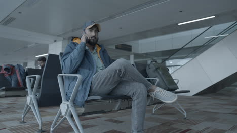 A-static-mid-shot-of-a-young-Indian-man-wearing-a-cap-and-a-jacket,-sitting-on-a-chair-at-an-airport,-talking-over-his-cellphone