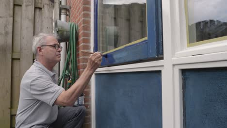 Reifer-Erwachsener-Mann,-Der-Einen-Fensterrahmen-Außen-Mit-Blauer-Farbe-Bemalt