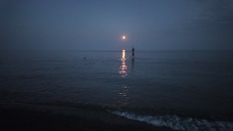 A-man-stands-on-a-paddleboard-and-rows-with-an-oar,-moving-along-the-path-of-the-moon's-reflection-in-the-water