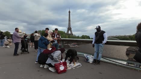 Tourists-negotiate-and-choose-from-mini-Eiffel-Tower-souvenirs-with-the-real-one-in-the-background-in-the-center-of-Paris,-France