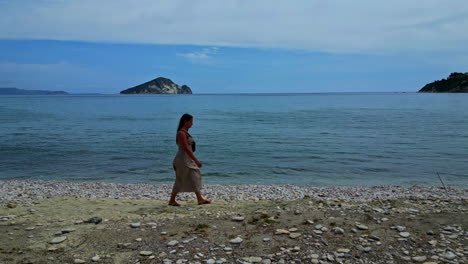 Mujer-Turista-Caminando-En-La-Orilla-Rocosa-De-Una-Playa-En-Grecia---Toma-Amplia