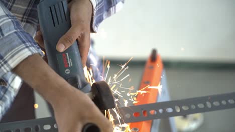 South-Asian-man-wearing-safety-goggles-working-with-an-angle-grinder-to-cut-slotted-angels-at-his-workspace