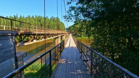 POV-shot-of-walking-forward-on-a-wooden-walkaway-bridge-over-a-river-beside-a-metal-bridge-during-daytime