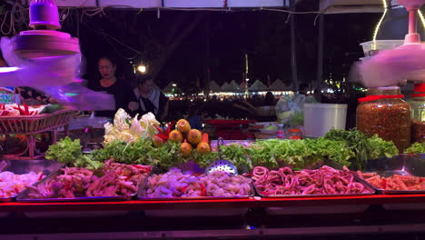 Abanicos-Con-Papel-Matamoscas-De-Plástico-Giran-Sobre-Bandejas-De-Verduras-Y-Mariscos-En-Un-Puesto-De-Un-Mercado-Nocturno-Asiático,-Plano-General