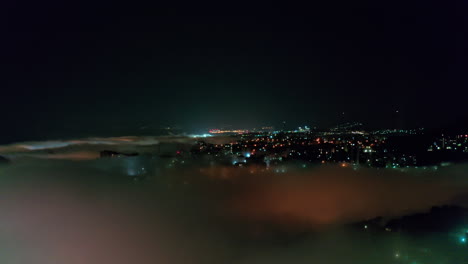 Vista-Aérea-Nocturna-De-Un-Paisaje-Urbano-Con-Edificios-Que-Emergen-A-Través-De-Una-Densa-Capa-De-Niebla.