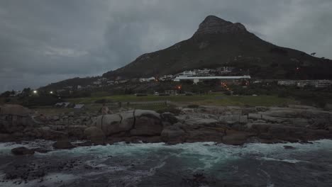 Drone-flying-backwards-over-the-sea-at-Camps-Bay-beach-in-Cape-Town-South-Africa---It's-cloudy-and-the-waves-are-crashing-against-rocks-with-a-view-of-the-Lion's-Head-mountain