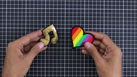 Hands-holding-glittery-music-note-patch-and-colorful-rainbow-heart-patch-against-a-grid-background