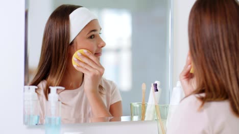 beauty,-hygiene-and-people-concept--teenage-girl-with-cleansing-sponge-cleaning-facial-skin-and-looking-in-mirror-at-bathroom