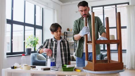 repair,-diy-and-home-improvement-concept--father-and-son-in-protective-gloves-stirring-grey-color-paint-in-can-for-painting-old-wooden-table-at-home