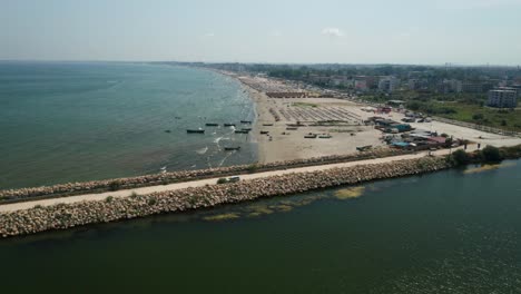 A-coastal-town-with-a-beach,-boats,-and-a-long-pier-on-a-sunny-day,-aerial-view